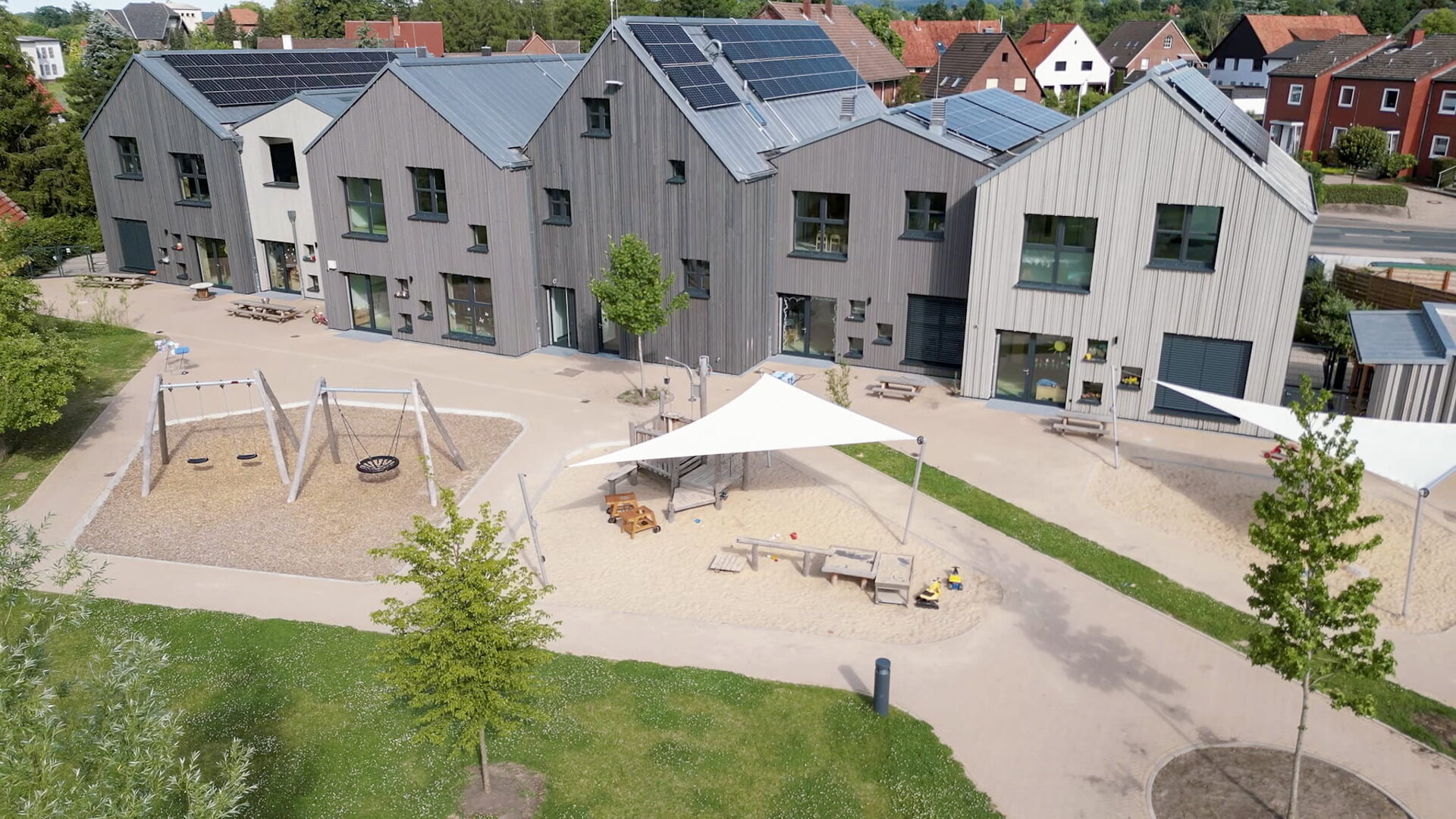 Ein modernes Kindertagesstättengebäude mit grauer Holzfassade, Solarmodulen auf dem Dach und einem Außenspielplatz mit Schaukeln, Sandkasten, Bäumen und einem schattigen Sitzbereich – fachmännisch errichtet vom Generalunternehmen Gundlach aus Hannover – umgeben von einem gepflasterten Gehweg.